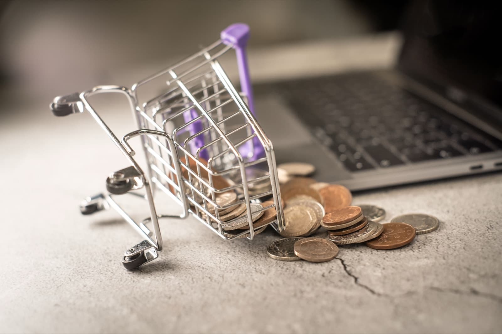 Miniature metal shopping cart spilling coins beside an open laptop, symbolising lost revenue from abandoned online carts