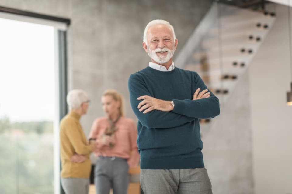 A smiling senior man with arms crossed standing in a modern office with two colleagues conversing near a staircase, representing an established firm principal