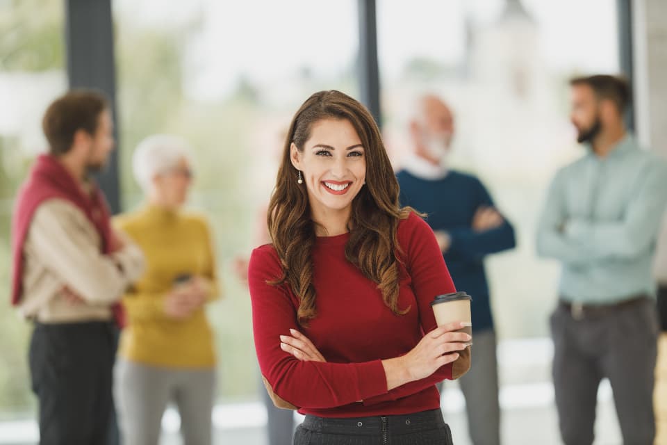A confident professional woman holding a coffee cup smiling in a modern glass-walled office with colleagues talking in the background