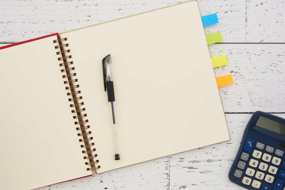 Overhead flat-lay of an open spiral notebook with a pen, a calculator and colored index tabs on a white wood desk, representing bookkeeping and planning