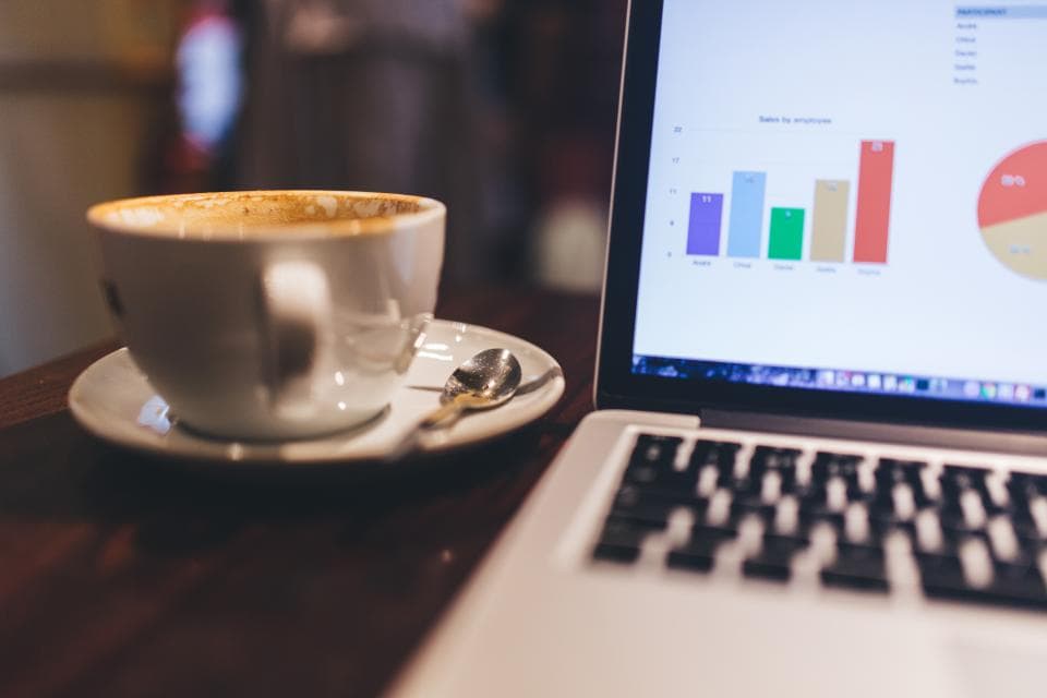 Laptop showing a coloured bar chart of sales by employee next to a cup of coffee on a dark wooden cafe table