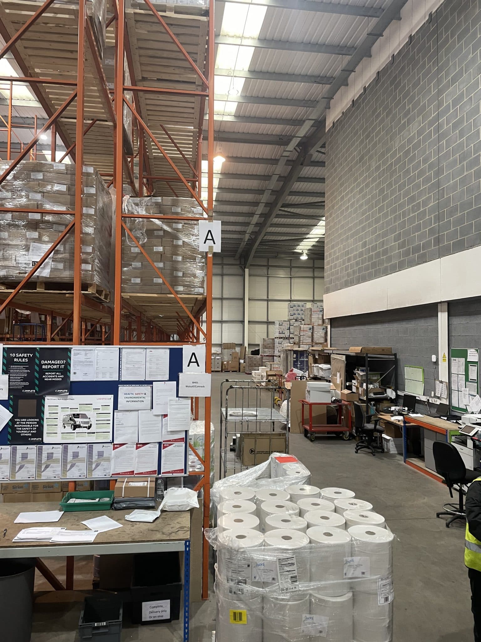 Interior of a working warehouse with tall orange pallet racking, wrapped stock, packing bench and a worker in a hi-vis vest