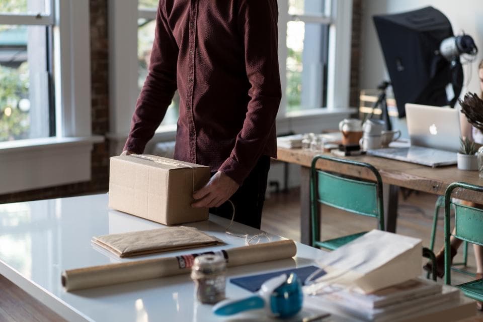 Person in a maroon shirt sealing a small cardboard parcel on a white packing table in a bright studio workspace
