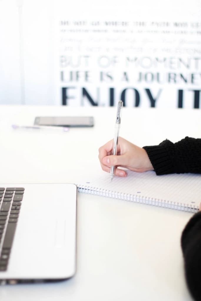 Person in a black jumper writing in a spiral notebook beside a laptop at a bright white desk
