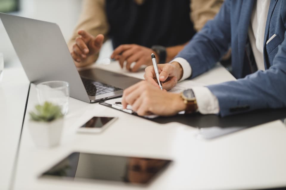 Two people at a desk reviewing documents beside a laptop, phone and small plant during a business meeting
