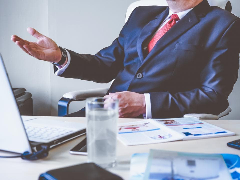 Business owner in a suit gesturing while seated at a desk with an open laptop, water glass and brochure