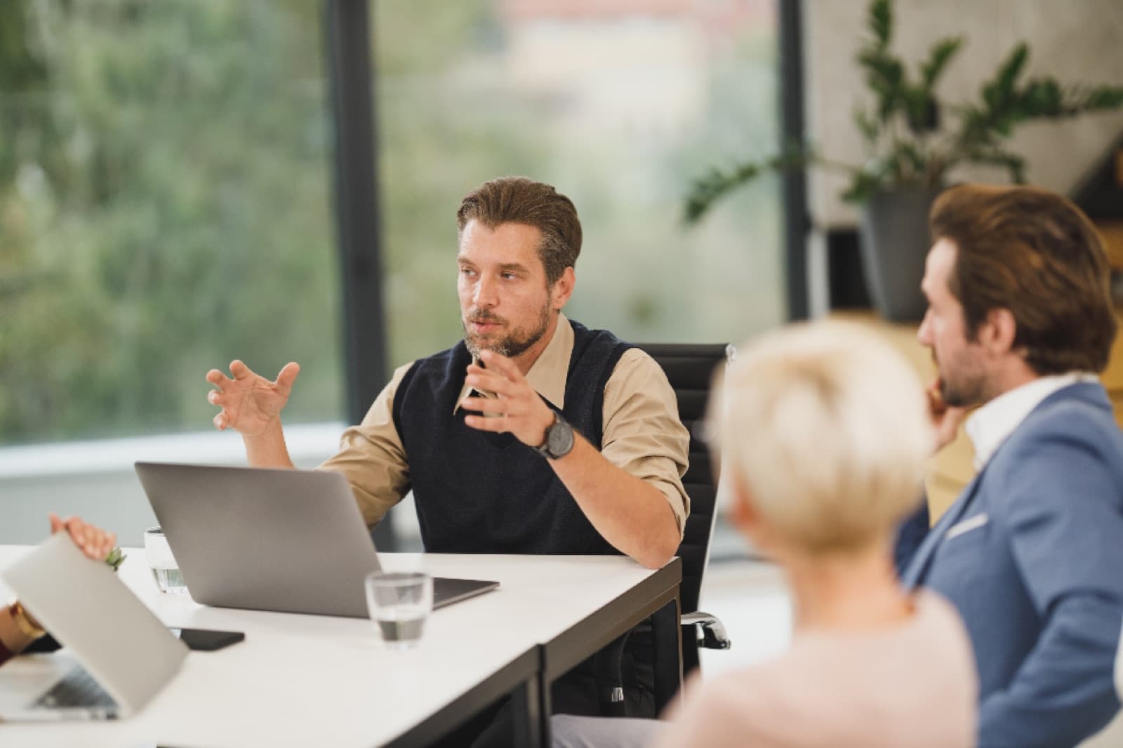 Man gesturing while talking in a bright modern office meeting with colleagues and laptops on a white table