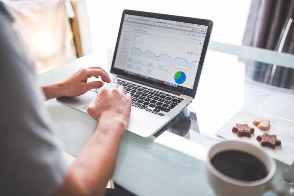Person typing on a laptop that shows a web analytics dashboard with a line graph and pie chart, coffee and biscuits on the