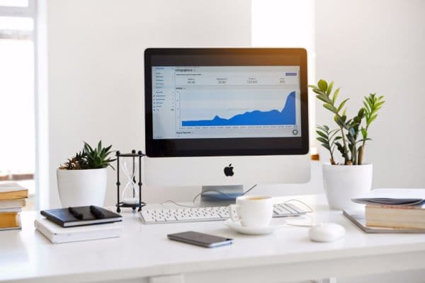 Desktop monitor on a tidy white desk showing an analytics dashboard with an upward trend graph, beside plants and coffee
