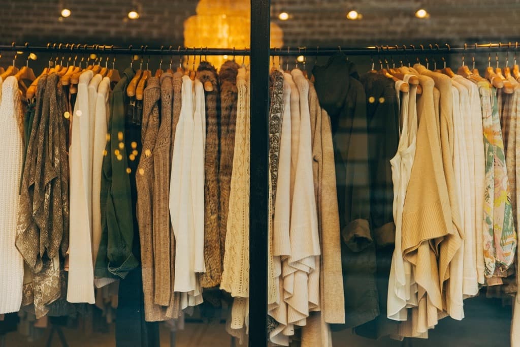 Rows of knitwear, cardigans and sweaters on wooden hangers seen through the window of a warmly lit clothing store