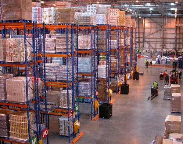 Large warehouse with tall pallet racking stacked with packed goods and a worker moving a pallet jack