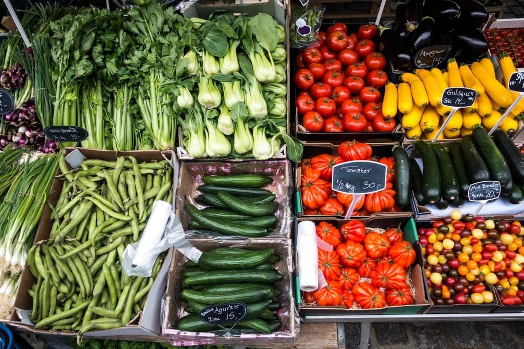 Fresh vegetables sorted into boxes at a grocery market, tomatoes cucumbers and greens each with handwritten price cards