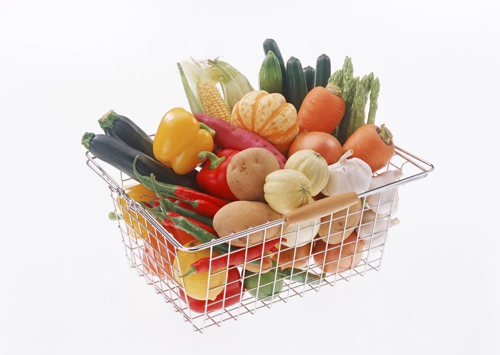 A wire grocery shopping basket filled with fresh vegetables including peppers corn onions and garlic on white