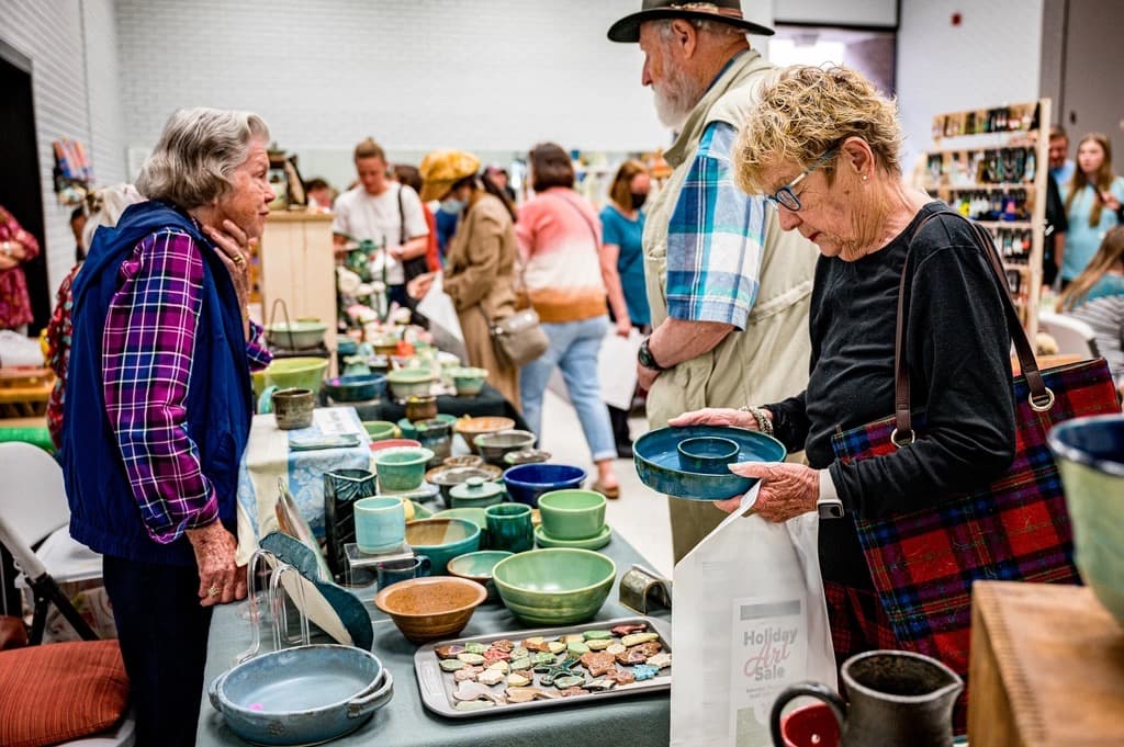 Shoppers browsing handmade ceramic bowls and mugs at a busy craft market stall while a seller talks with a customer