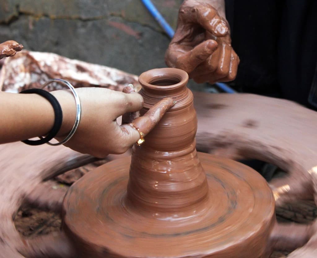 Two pairs of hands, one wearing glass bangles, shaping a wet terracotta pot on a spinning potter's wheel in a workshop