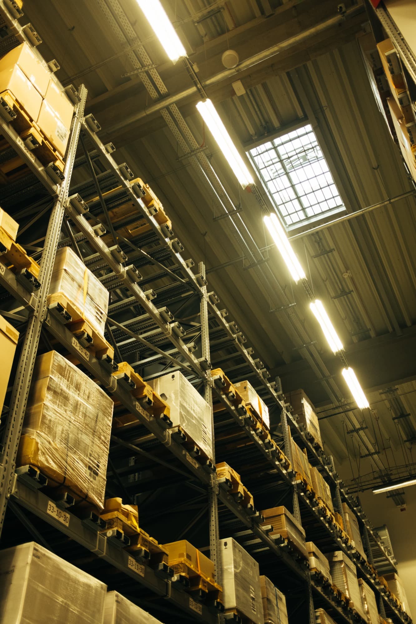 Tall warehouse racking filled with palletised, shrink-wrapped boxes under industrial lighting
