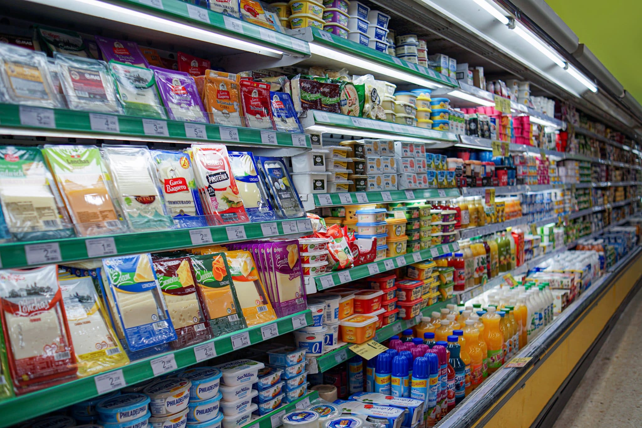 Densely stocked refrigerated grocery aisle with packaged cheese, dairy, spreads and bottled drinks