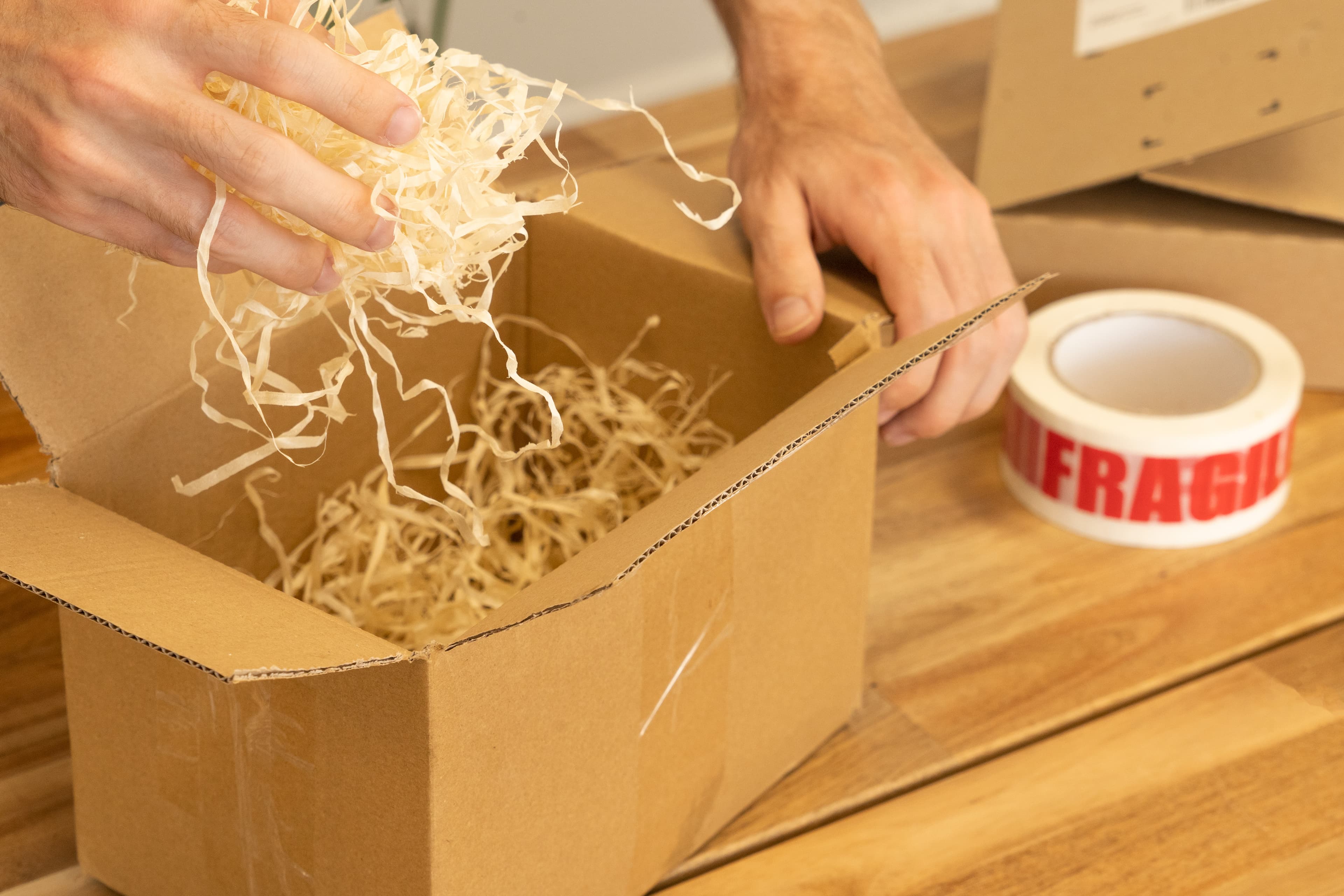 Hands filling a cardboard parcel with paper wood wool next to a roll of fragile tape for a customer order