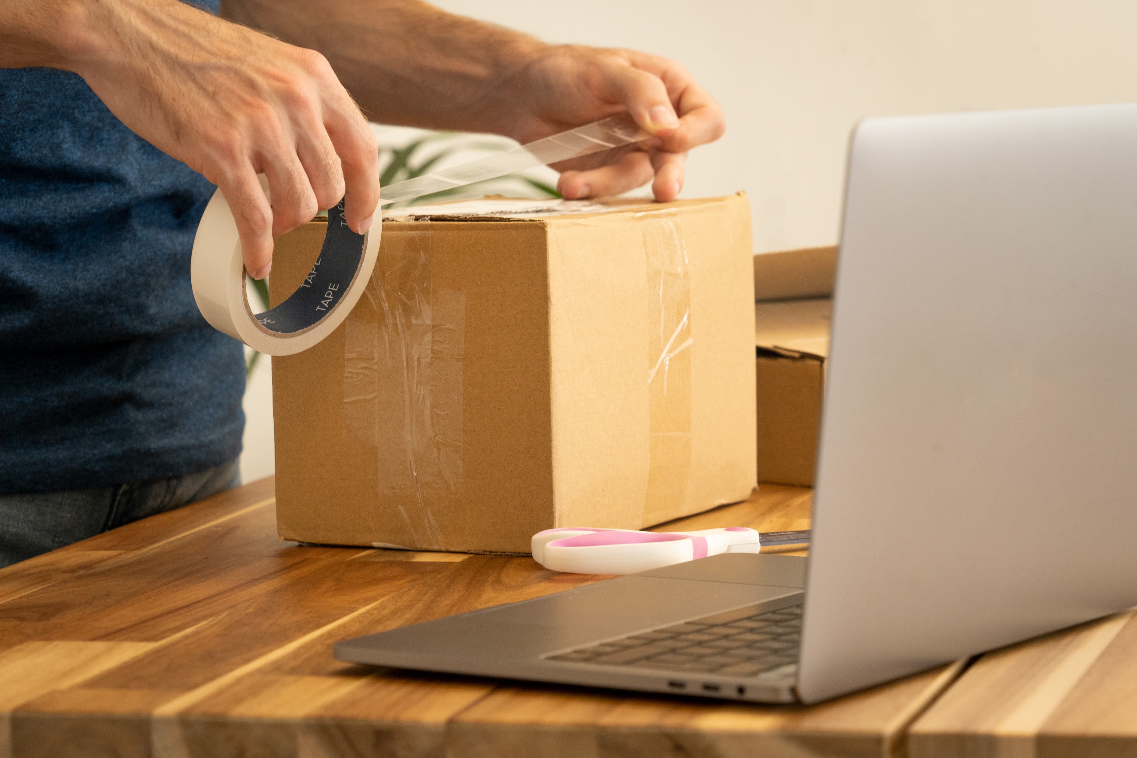 Online seller taping a cardboard shipping box with a laptop open on the desk beside packing supplies