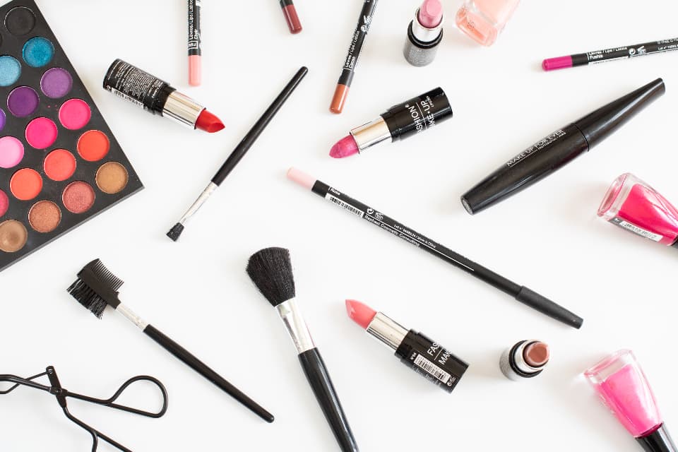 Flat-lay of makeup products including an eyeshadow palette, lipsticks, brushes, liners and nail polish on white