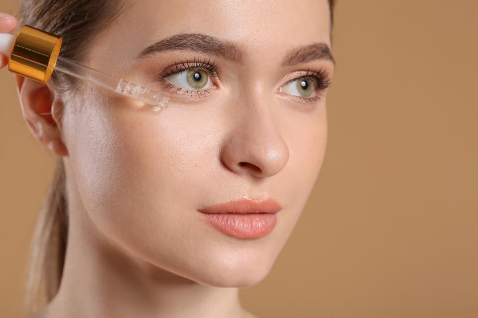 Woman applying a facial serum from a glass dropper to glowing skin against a warm beige studio background