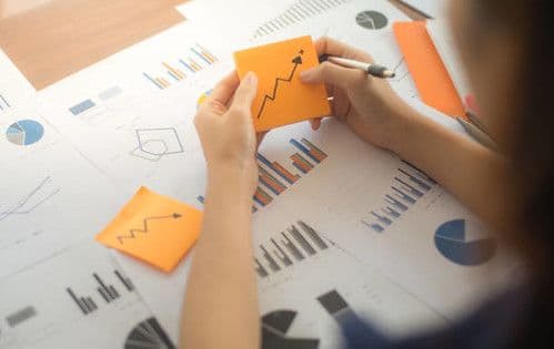 Hands arranging orange sticky notes showing upward growth arrows over printed sales charts and analytics reports on a desk
