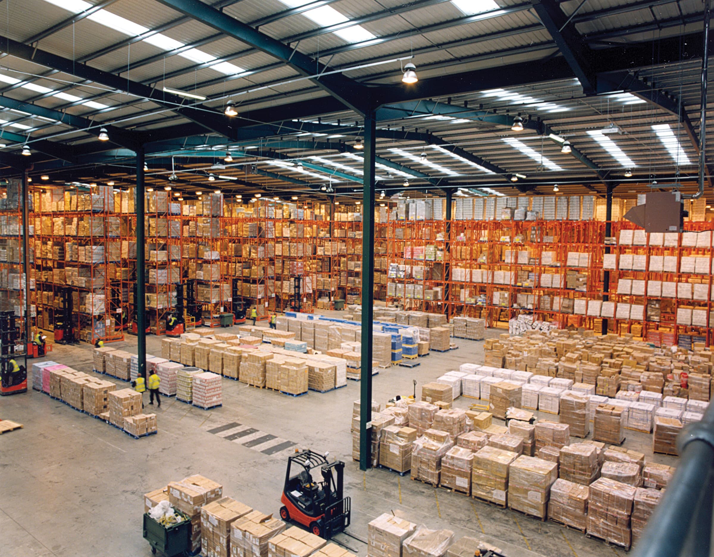 Interior of a busy distribution warehouse with tall orange pallet racking stacked with boxes and forklifts moving orders