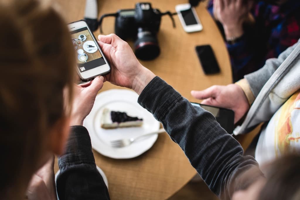 Person holding a smartphone to photograph a plated dessert at a cafe table beside a camera