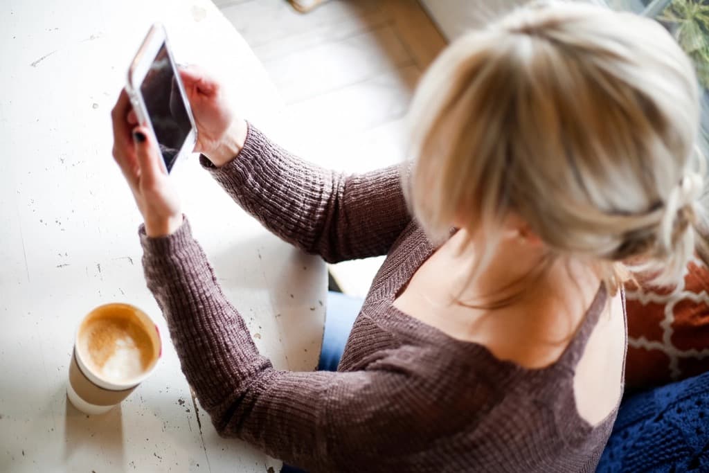 Overhead view of a woman holding a smartphone at a white table with a cup of coffee