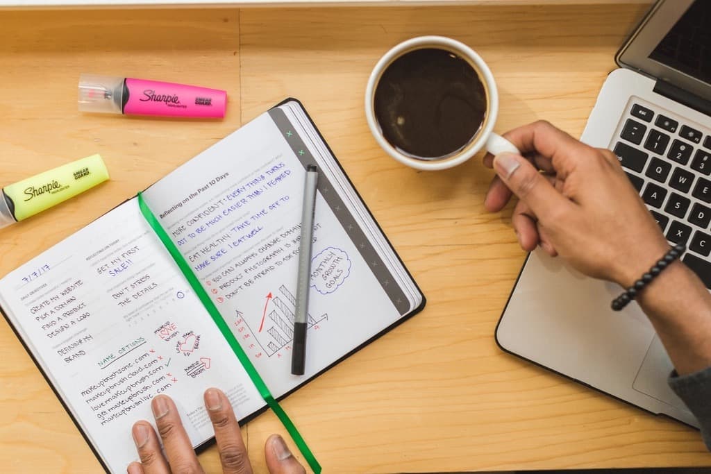 Open notebook with handwritten plans to create a website and make a first sale, beside a coffee cup and laptop