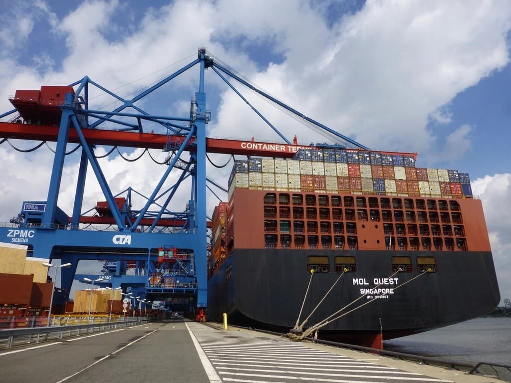 Large container ship docked at a port container terminal beneath blue ship-to-shore gantry cranes, stacked with multicolored shipping containers