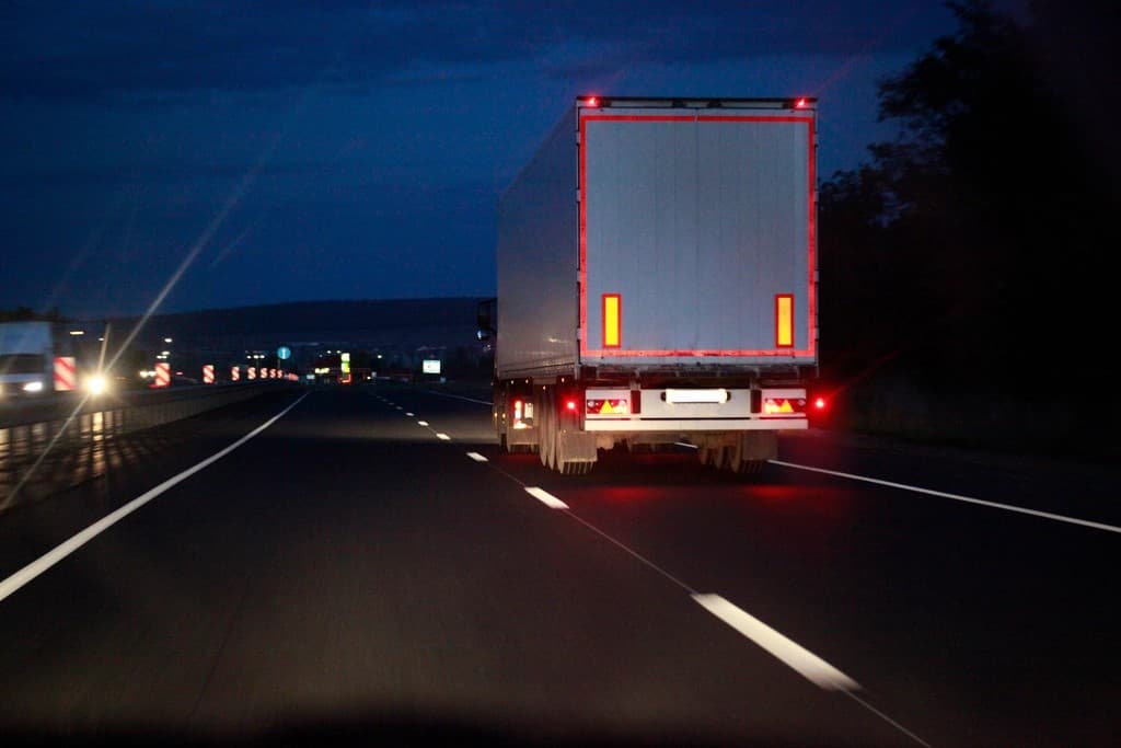 Rear view of a semi-trailer freight truck driving on a highway at dusk with glowing red tail lights and reflective markers