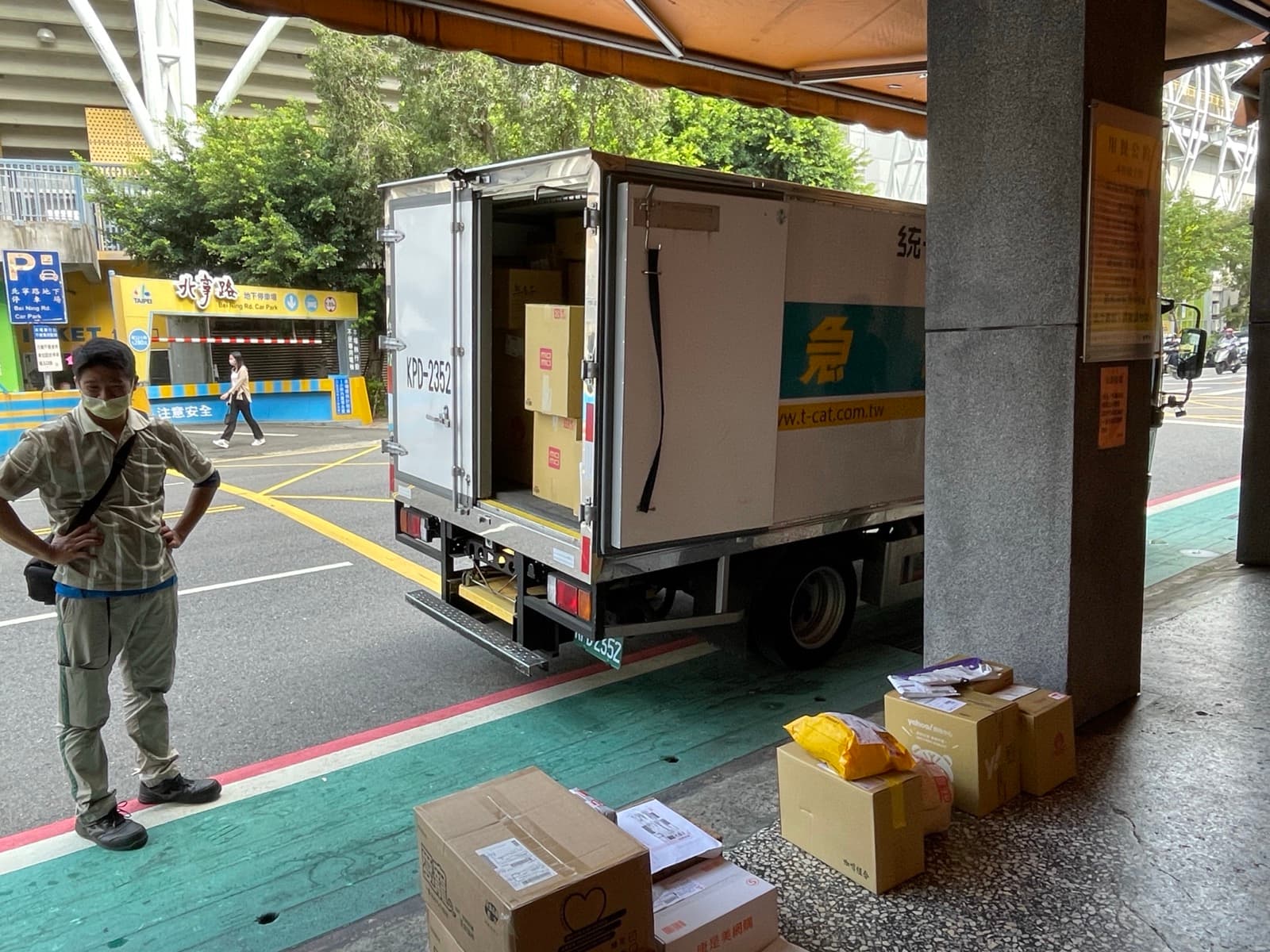 Courier standing beside an open delivery van on a city curb with cardboard parcels staged on the sidewalk for last-mile drop-off