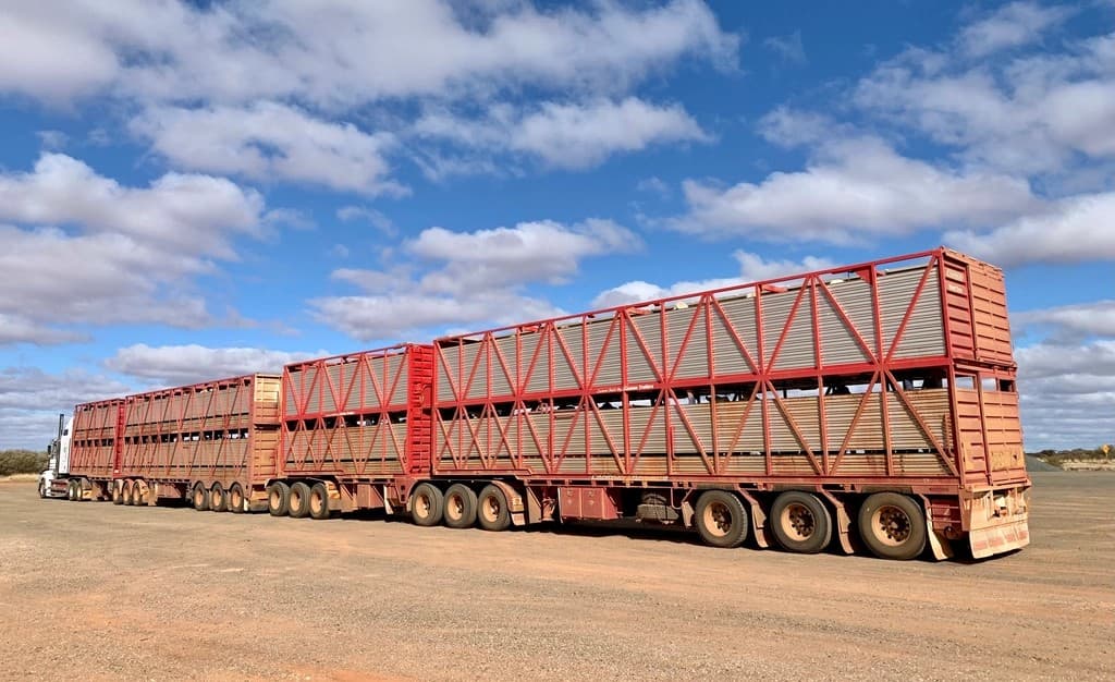 Multi-trailer road train freight truck parked on a dirt lot under a bright blue sky with scattered clouds