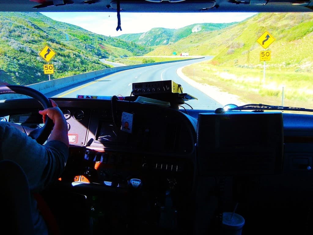 First-person view from inside a truck cab, driver's hand on the wheel and dashboard controls, looking out over a winding highway through green hills