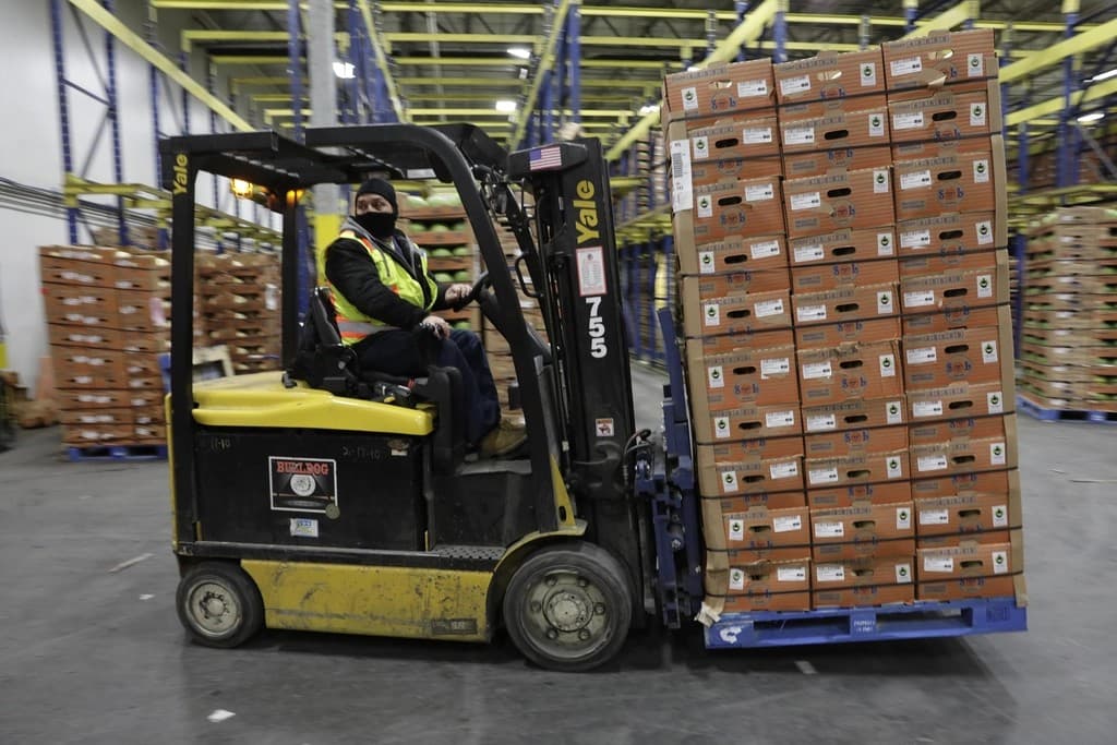 Warehouse worker in a hi-vis vest driving a forklift loaded with a stacked pallet of labeled cartons down a racked distribution-center aisle
