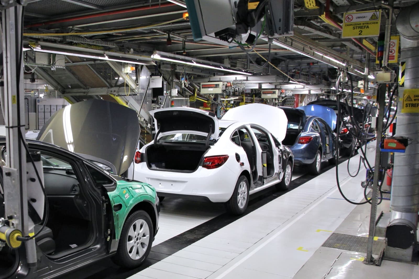 Automotive assembly line with car bodies moving on a conveyor, hoods and trunks open, overhead tooling and gantries in an auto plant