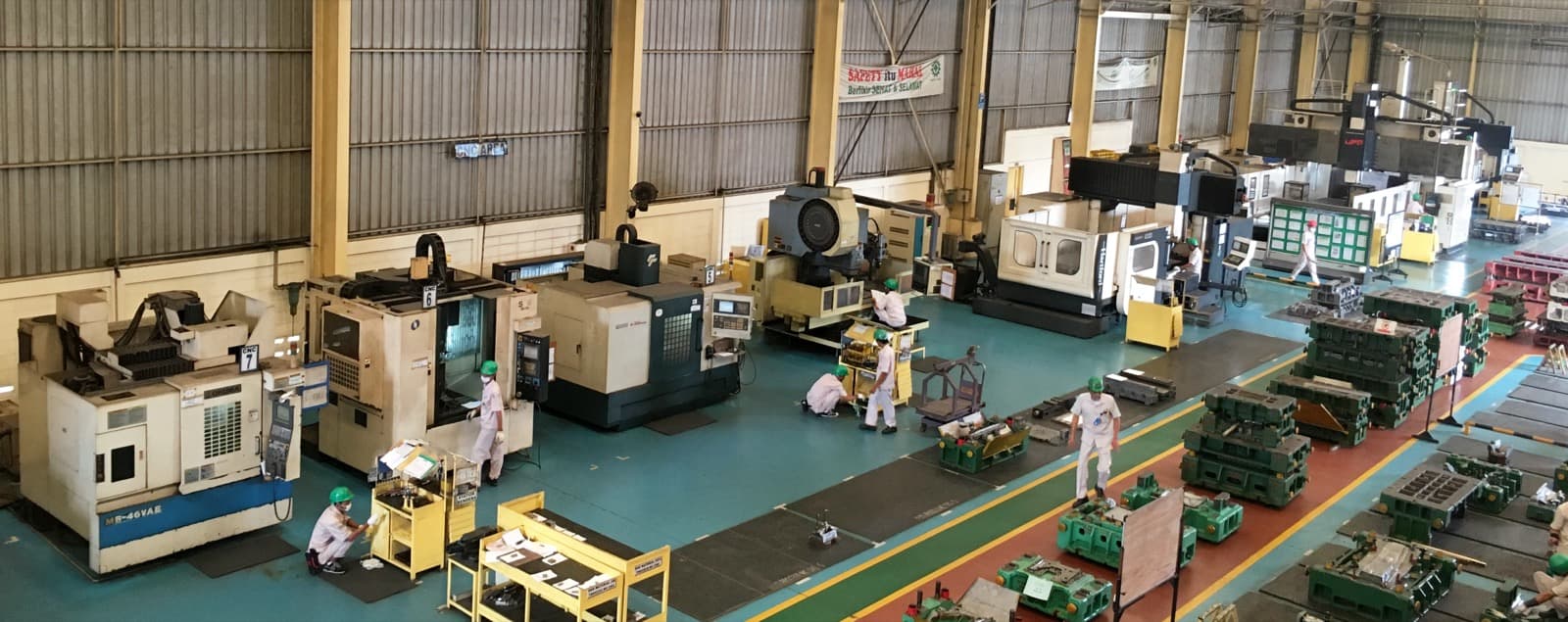 Overhead view of a CNC machine-shop floor with numbered machining centers, green metal castings staged in rows and workers in hard hats