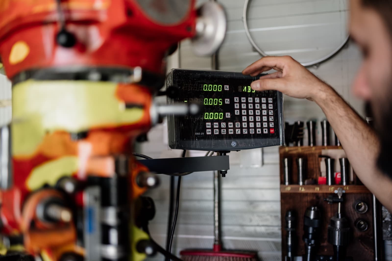 Machinist reaching to a green digital readout panel mounted on a milling machine in a metalworking shop, tool holders racked on the wall