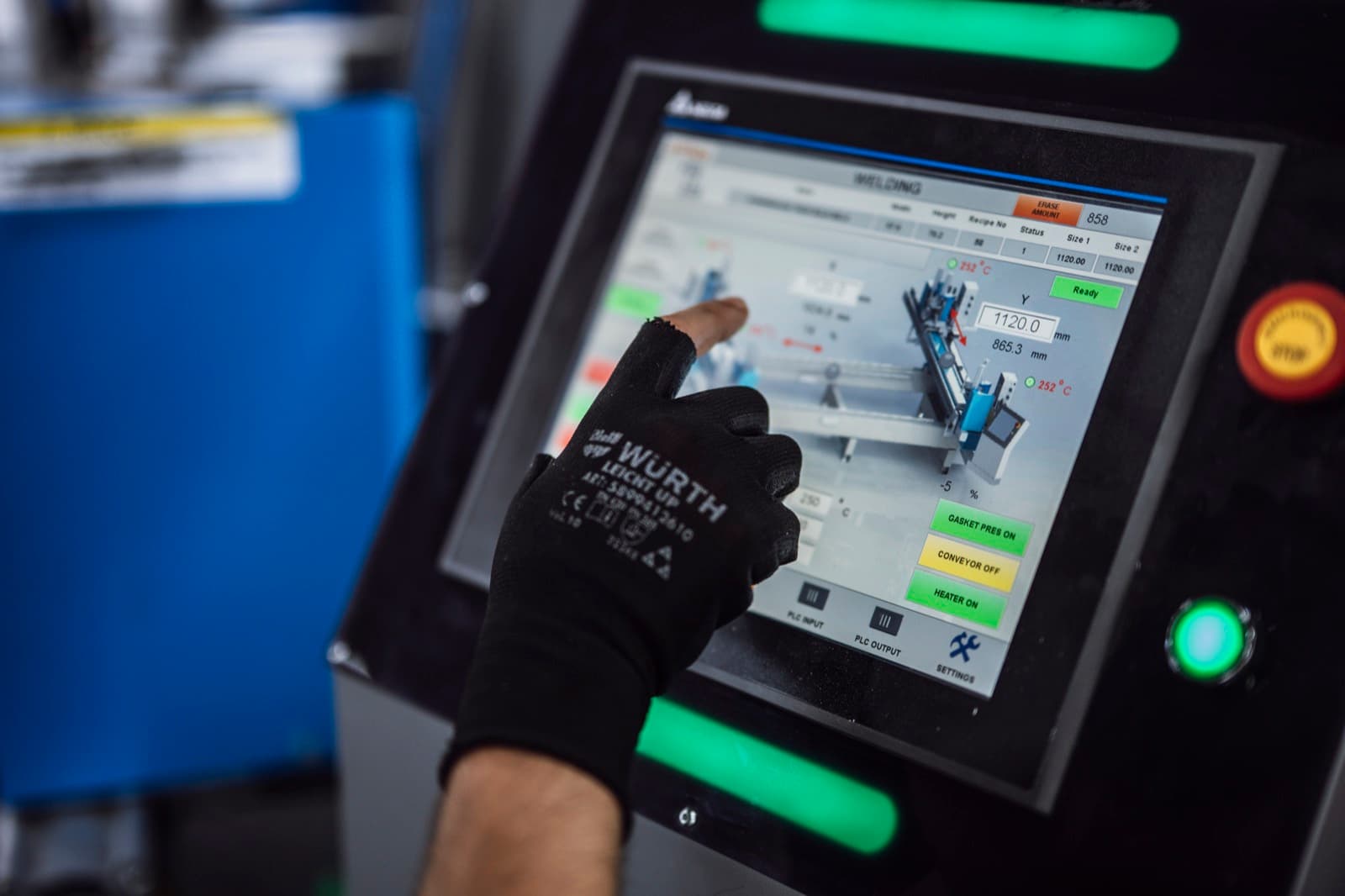 Factory operator in a work glove tapping an industrial HMI touchscreen showing a live machine control panel with temperature readouts and status flags
