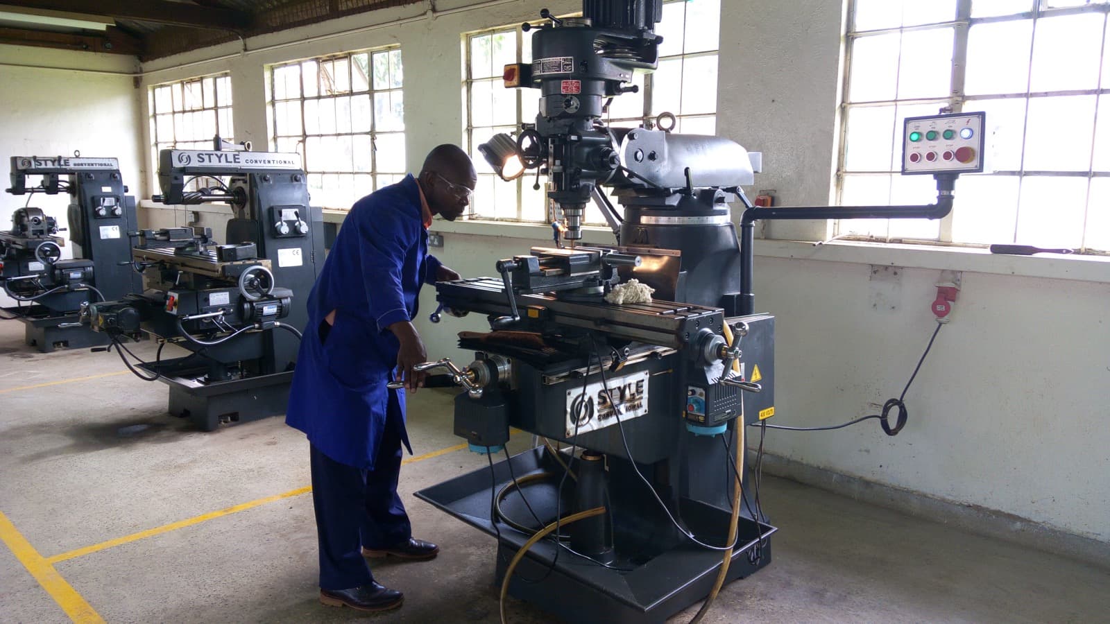 Operator in blue coveralls and safety glasses working a vertical milling machine on a machine-shop floor, additional machine tools in the background