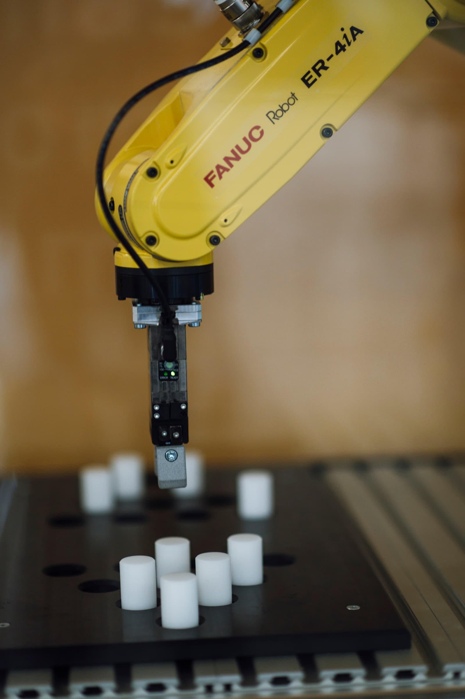 Yellow industrial robot arm with a gripper picking up white cylindrical parts from a fixture plate in an automated factory cell