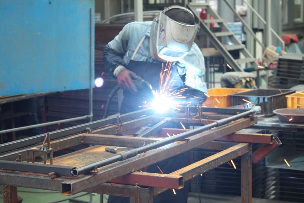 Welder in a face shield joining a steel frame with bright sparks flying in a metal-fabrication workshop